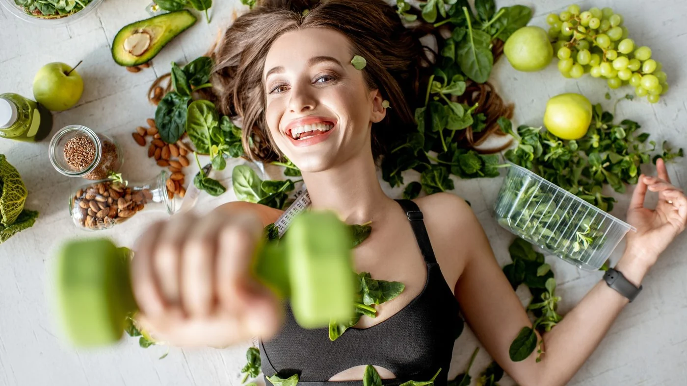 Девушка с гантелей в окружении зеленых продуктов